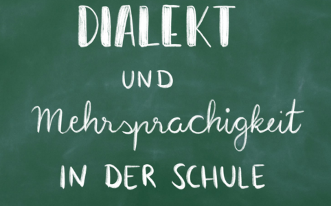 Auf dem Bild ist eine grüne Schultafel zu sehen, auf der mit weißer Kreide "Dialekt und Mehrsprachigkeit in der Schule" geschrieben steht.